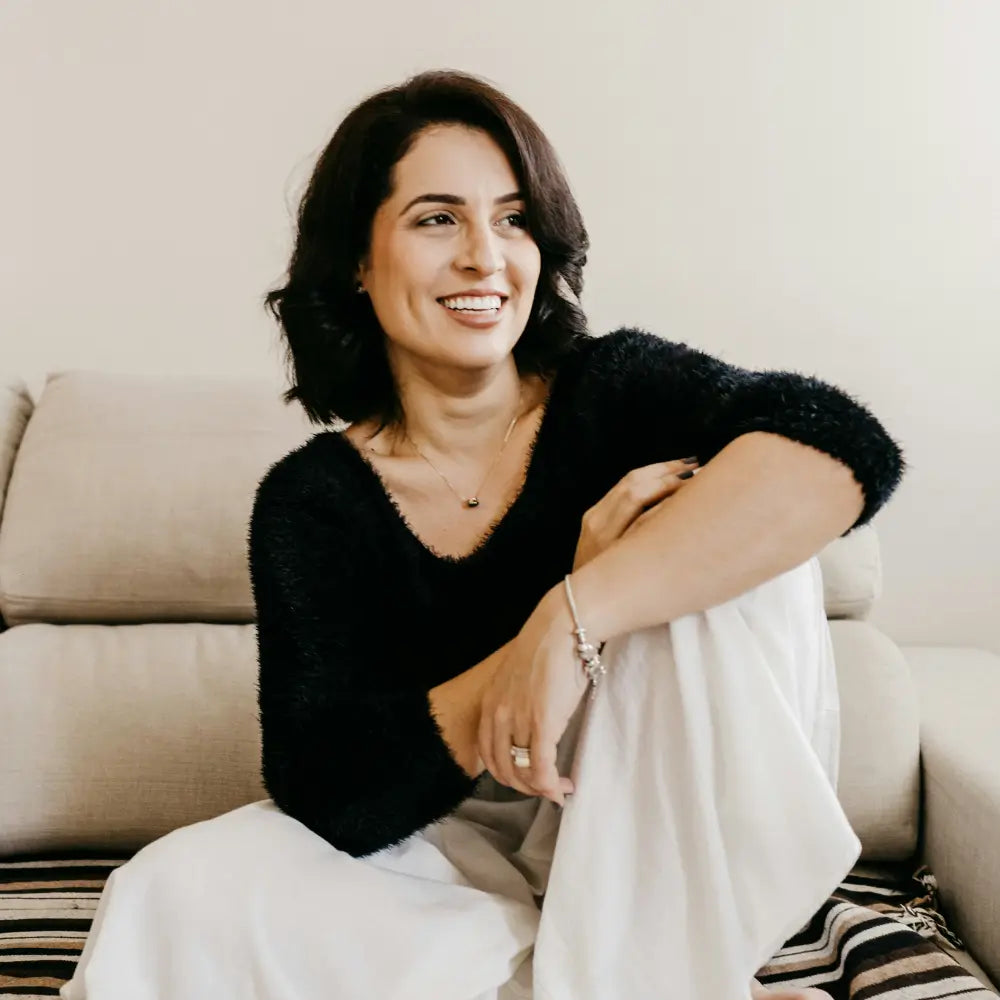 Woman sitting on a couch wearing a black sweater and white pants, smiling. Image credit Jonathan Borba